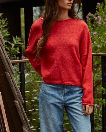 Woman wearing a red sweater and blue jeans standing on a wooden deck with greenery in the background