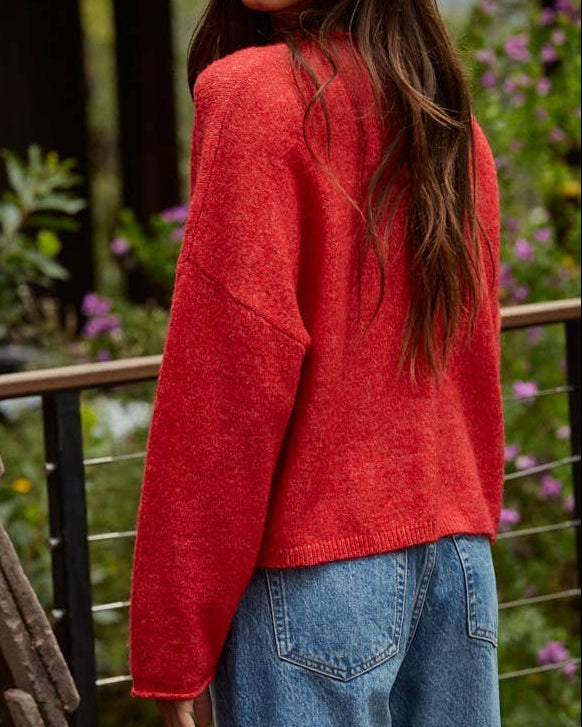 Woman wearing a red sweater and blue jeans standing outdoors with greenery in the background