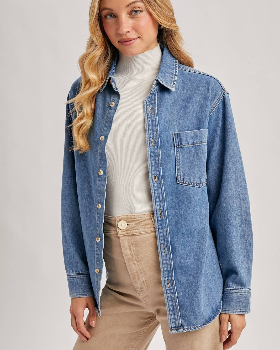 Woman wearing a denim shirt over a white sweater and beige pants on a light gray background