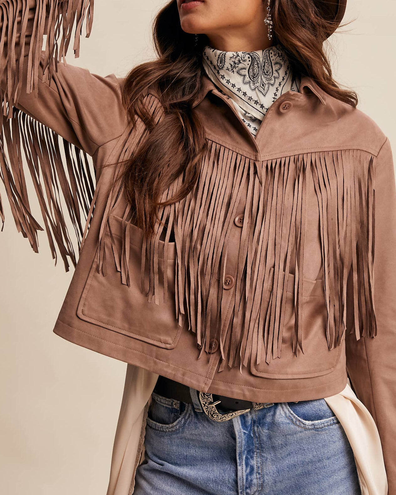 Woman wearing a brown fringed jacket and denim shorts against a beige background