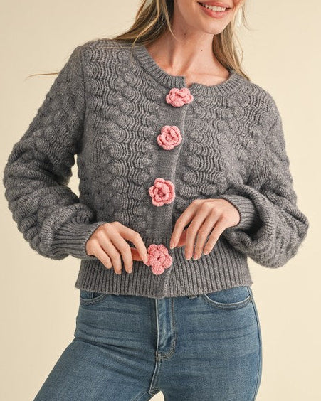 Woman wearing a gray cable knit cardigan with pink floral buttons on a beige background