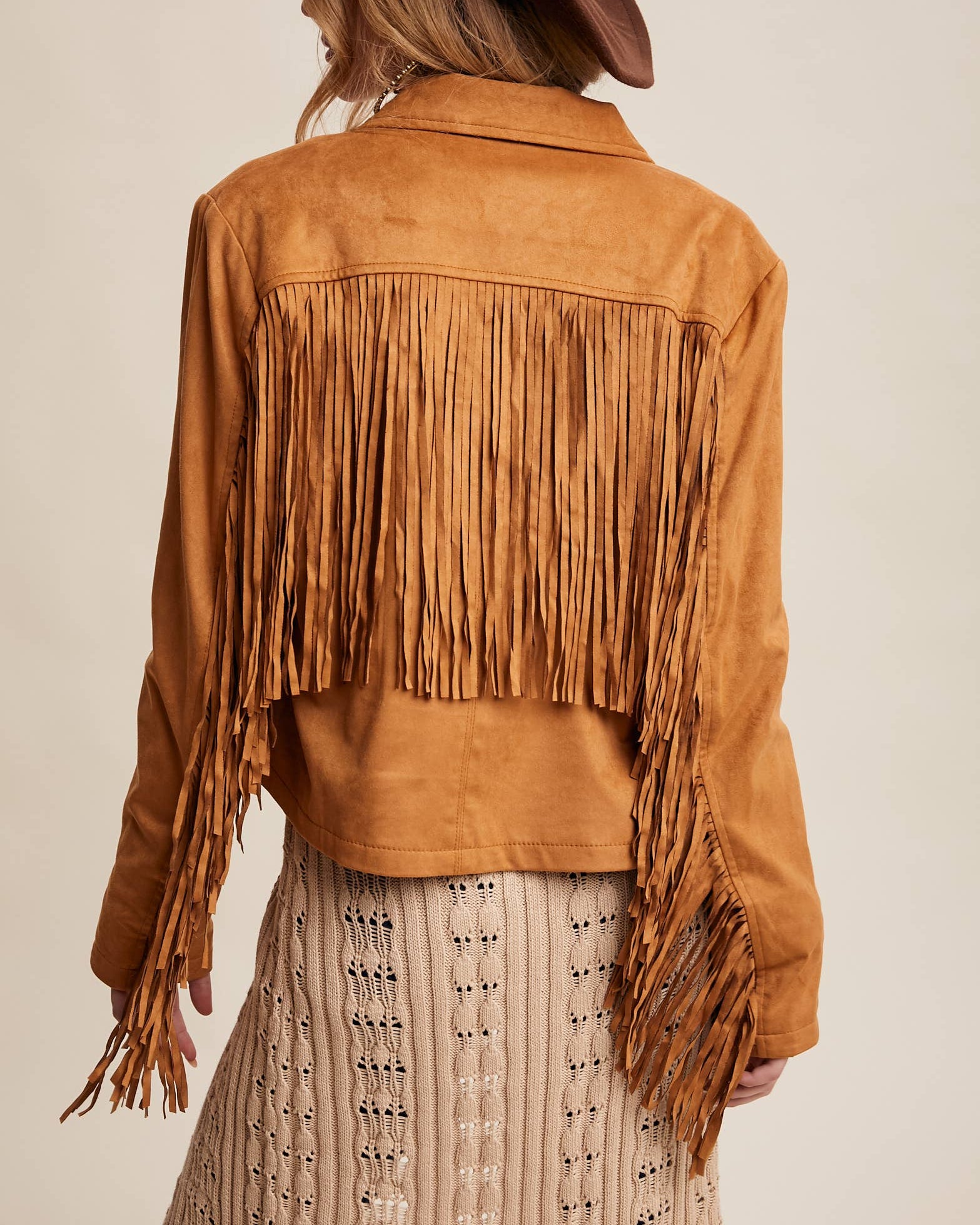 Person wearing a brown suede fringe jacket and hat on a beige background