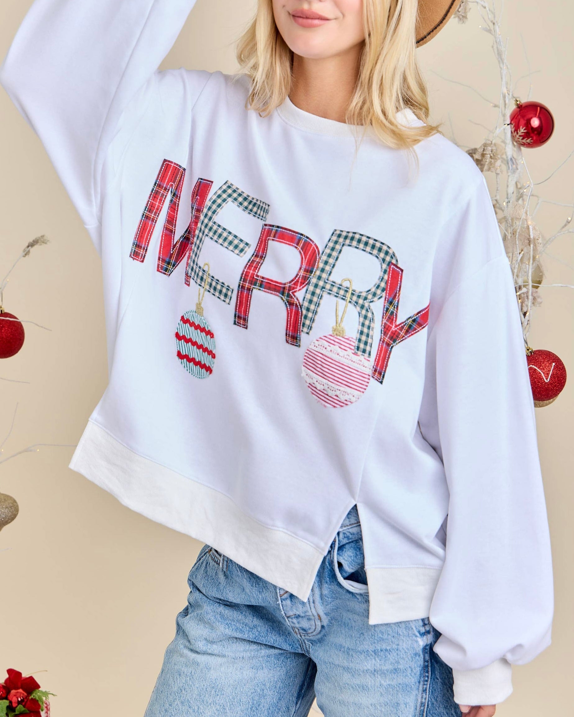 Person wearing a white sweatshirt with 'MERRY' text and Christmas-themed design, standing against a beige background with Christmas decorations.
