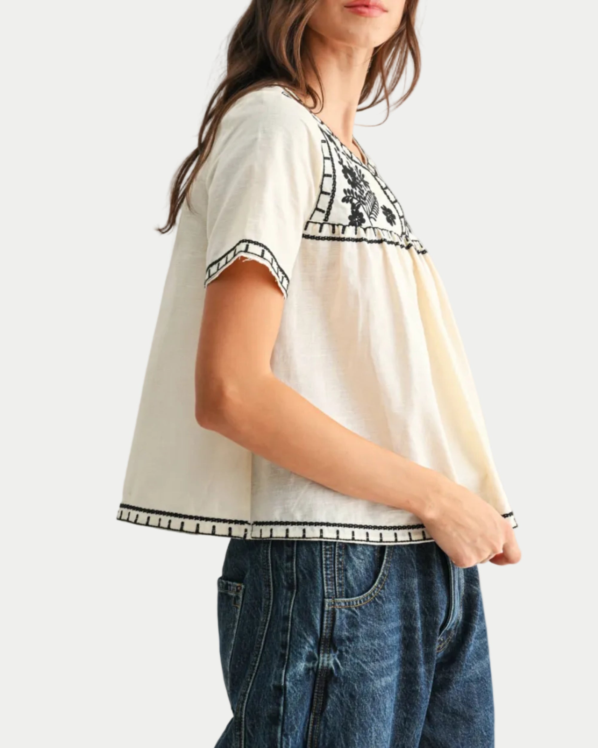 Woman wearing a white blouse with black embroidery and blue jeans on a white background side view