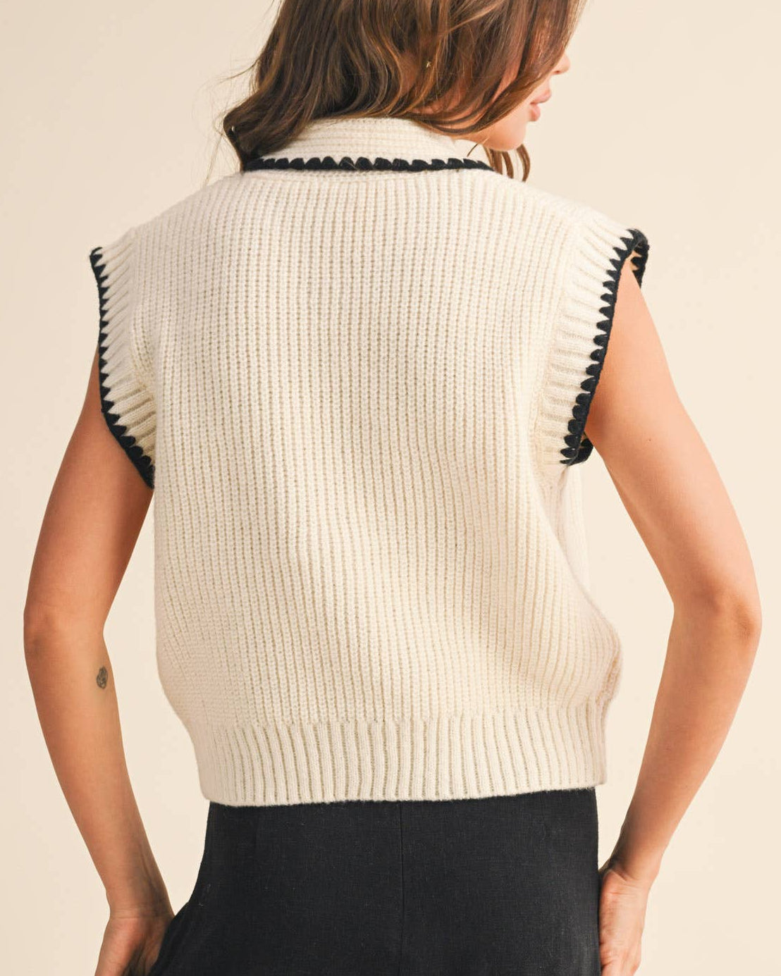 Woman wearing a cream-colored sleeveless sweater with black trim on a beige background