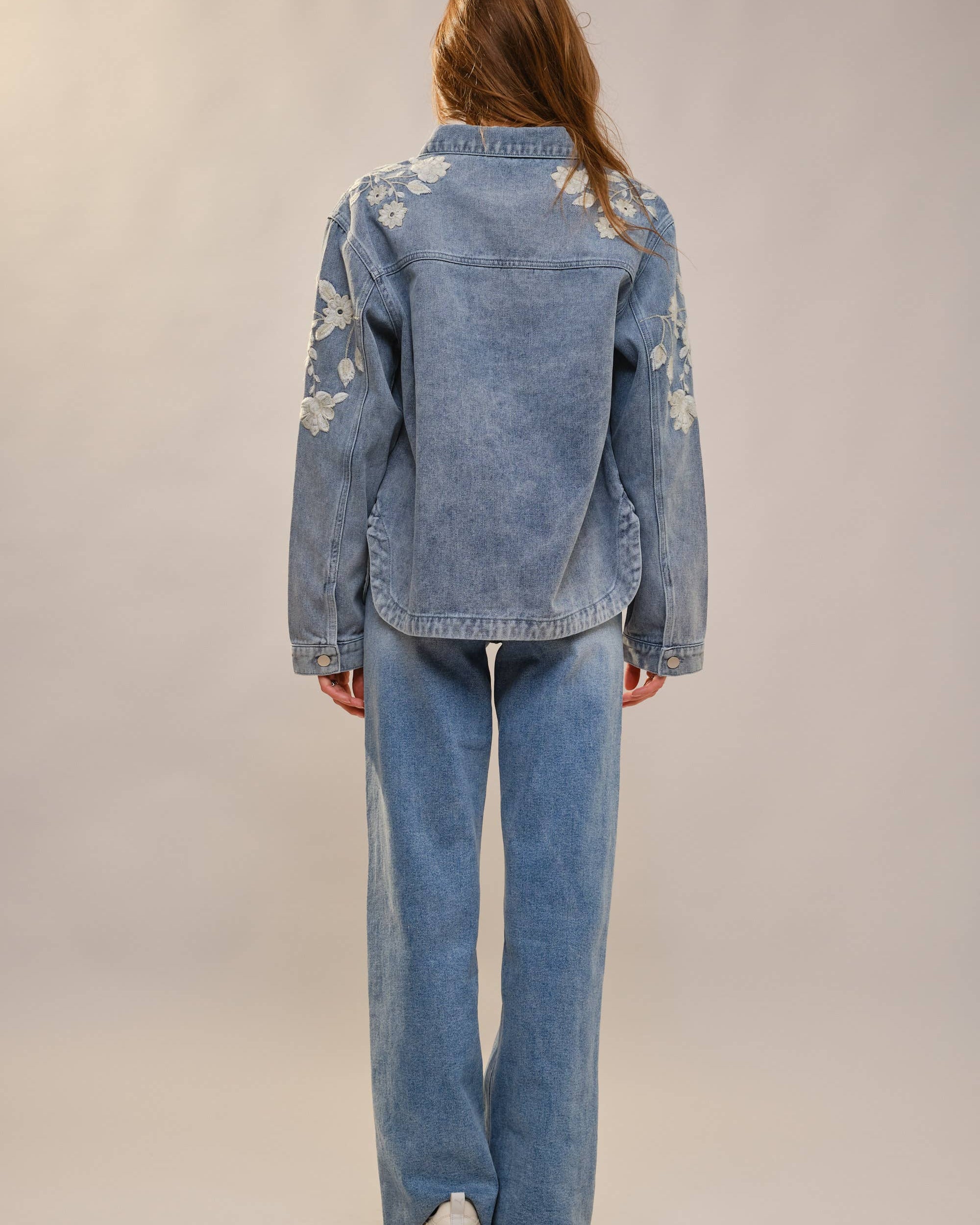 Person wearing a blue denim jacket with floral embroidery and matching jeans on a plain background