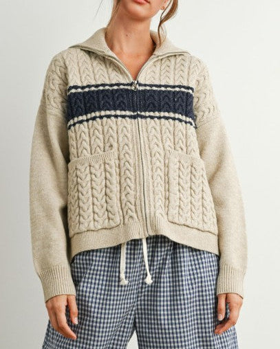 Woman wearing a beige knit hoodie with a navy stripe and plaid pants on a plain background