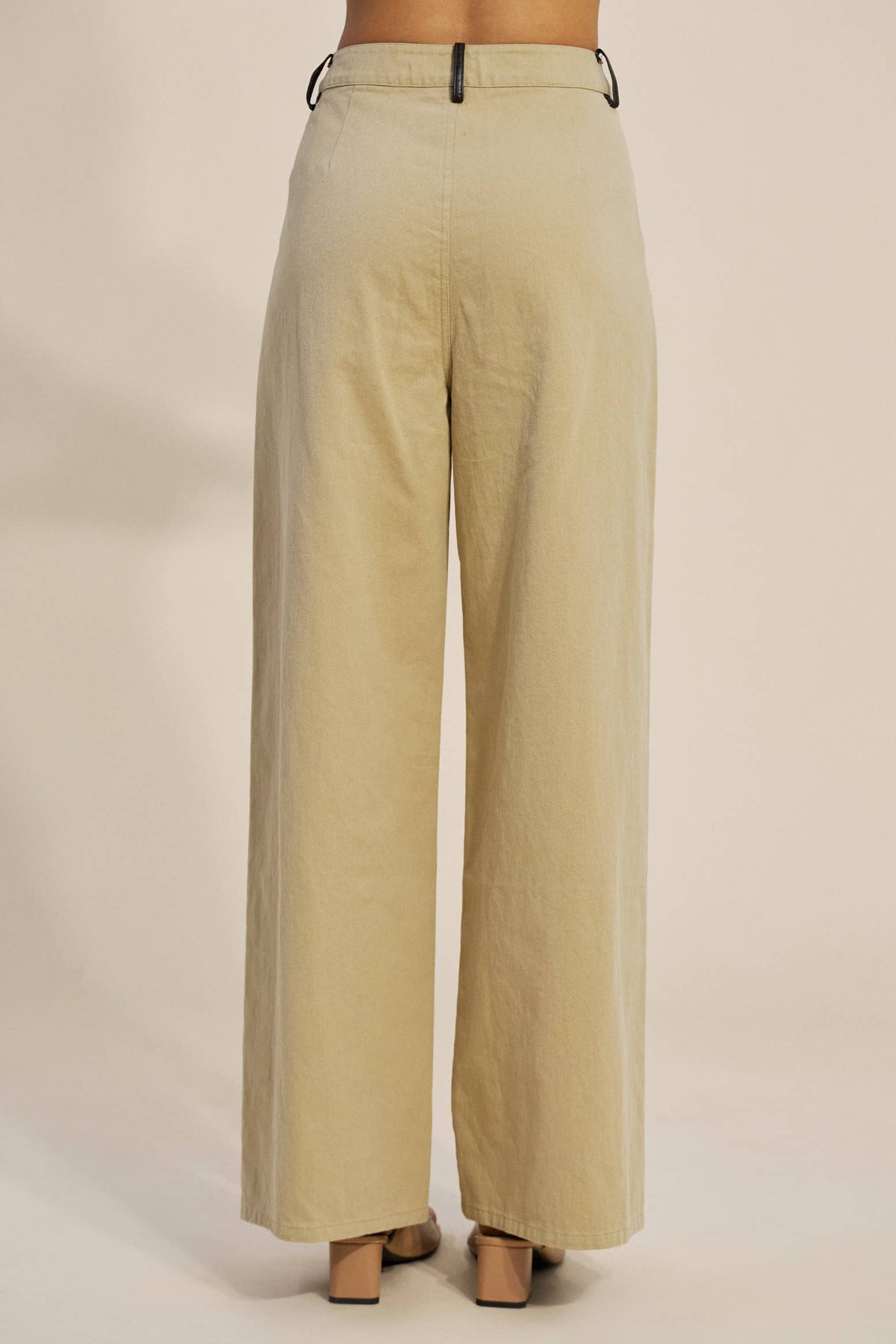 A person shown from the back wearing high-waisted, wide-leg trousers in a light beige or khaki color, paired with beige heels, against a plain beige studio background.