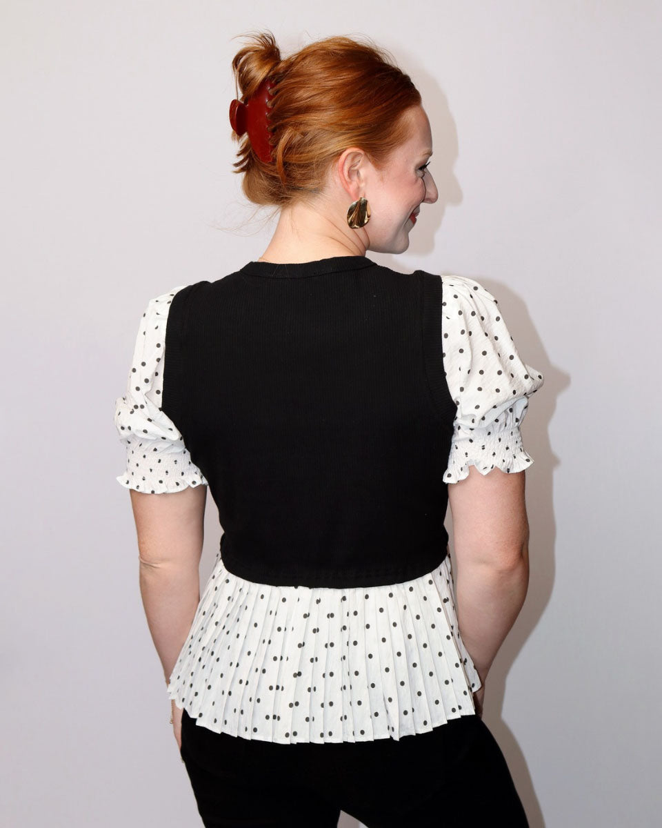 A woman with red hair tied up in a bun is shown from the back, wearing a black sleeveless vest over a white short-sleeve blouse with black polka dots and ruffled sleeves and hem.