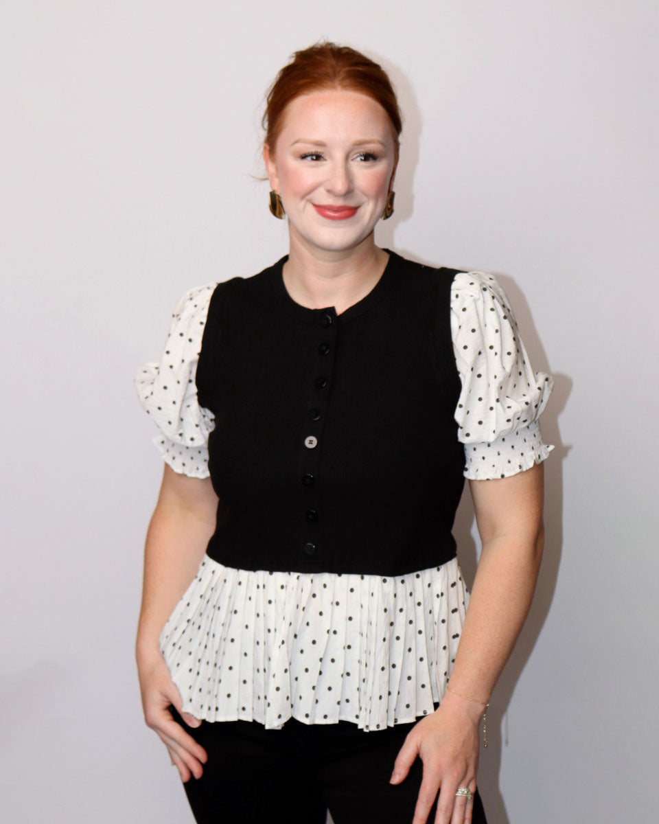 A woman with red hair tied back, wearing a black button-front vest over a white puff-sleeve top with black polka dots and a matching white pleated hem, standing against a plain light gray background.