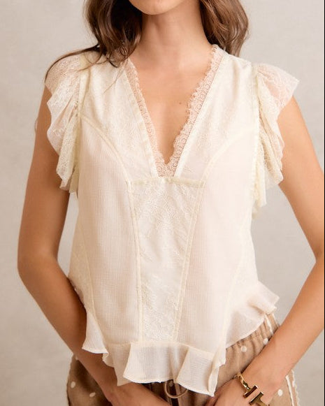 A Model wearing a cream-colored sleeveless blouse with ruffled flutter sleeves, a lace-trimmed V-neckline, and a ruffled hem on a neutral background