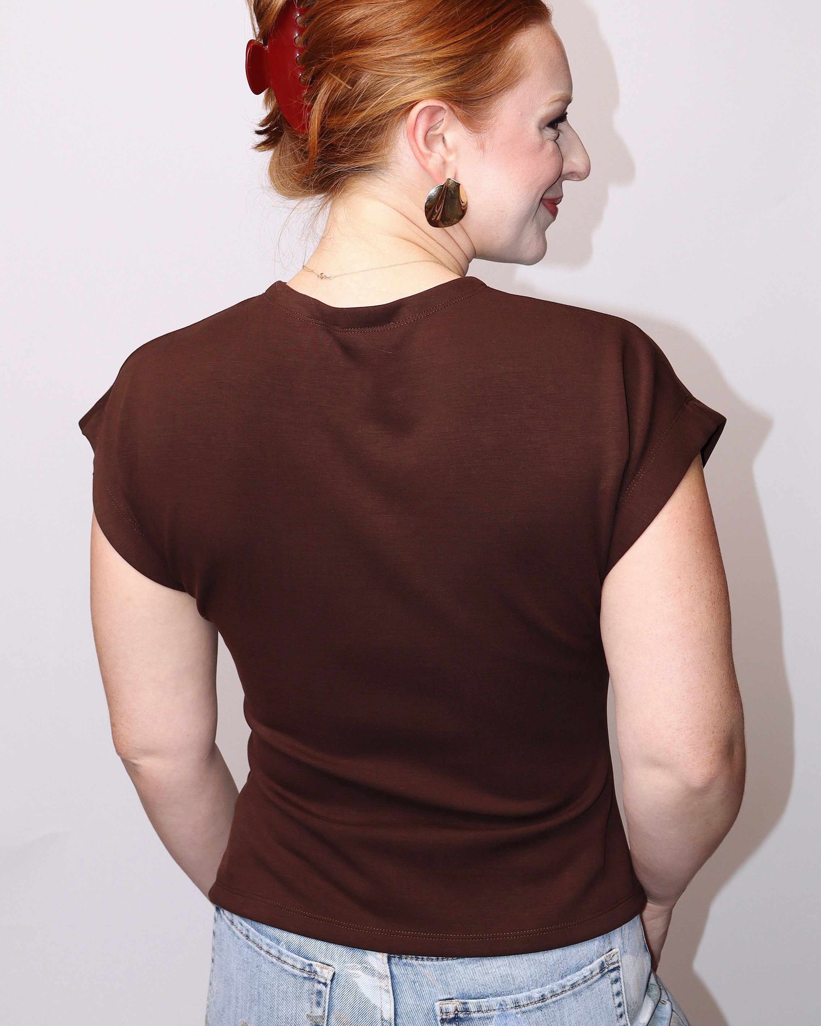 A woman with red hair tied up is shown from the back wearing a dark brown short-sleeve t-shirt and light blue jeans against a plain light gray background.
