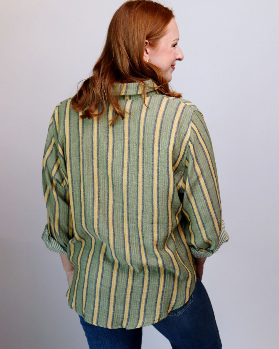 A woman with red hair is shown from the back wearing a green and yellow vertically striped button-up shirt and blue jeans against a plain light gray background.
