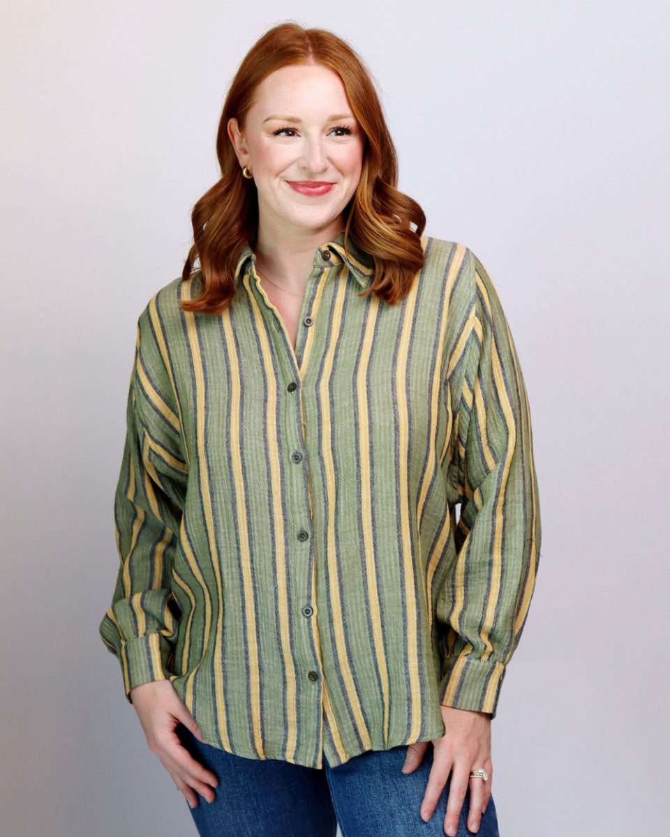 A woman with red hair wearing a green and yellow striped button-down shirt and blue jeans, standing against a light gray studio background.