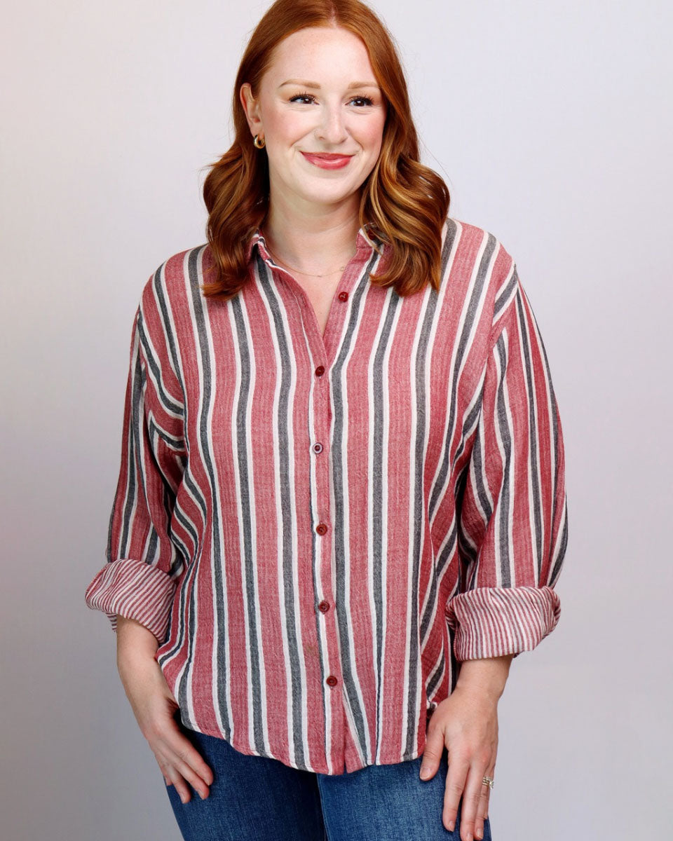 A woman with red hair wearing a red, white, and gray vertically striped button-down shirt with rolled-up sleeves, paired with blue jeans, against a light gray studio background.