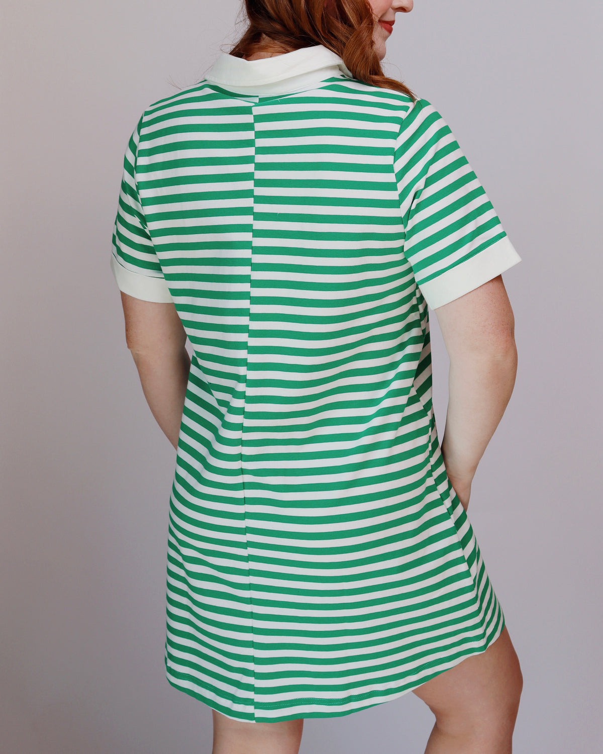 A woman wearing a green and white horizontally striped short-sleeve polo dress, shown from the back against a plain light gray studio background.