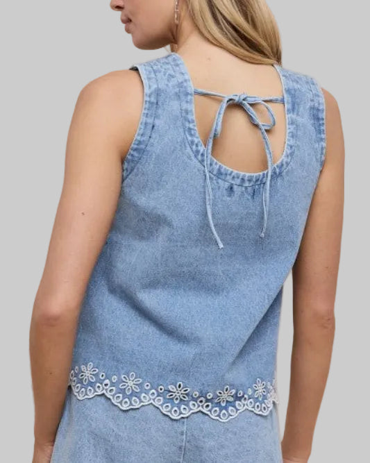 Model wearing a light blue sleeveless denim with eyelet embroidery at hem top on a neutral background. back view