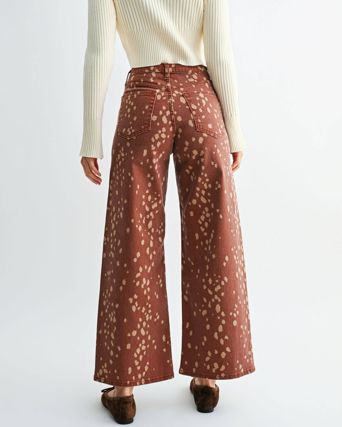 A woman shown from the back wearing a cream ribbed turtleneck sweater and rust-brown wide-leg pants with a small beige floral pattern, set against a plain light gray studio background. Bambi Deer Print Seamed Slim Wide Leg Jeans | Blackbird Boutique