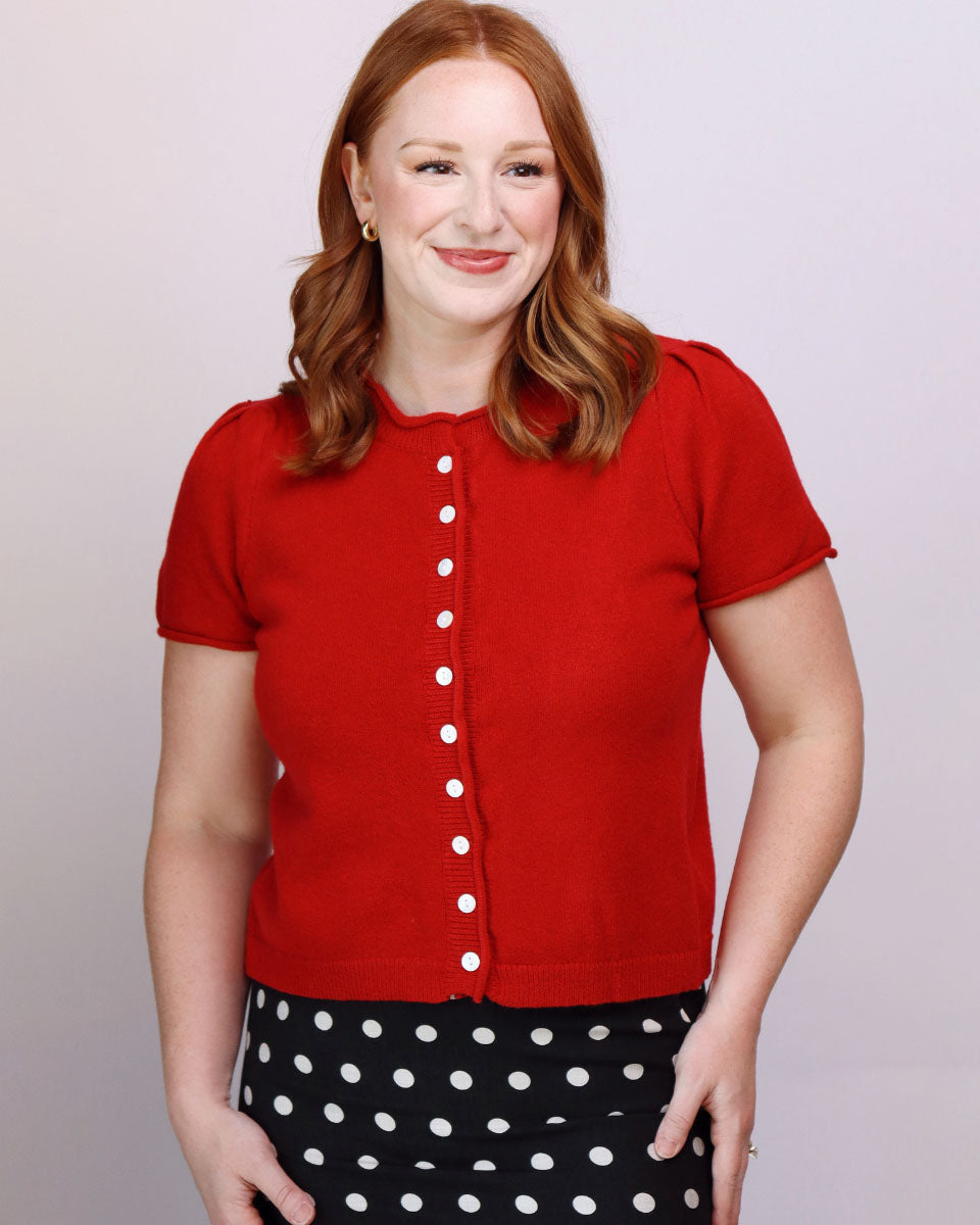 A woman with red hair wearing a red short-sleeve button-front top and a black-and-white polka dot skirt, standing against a light gray studio background.