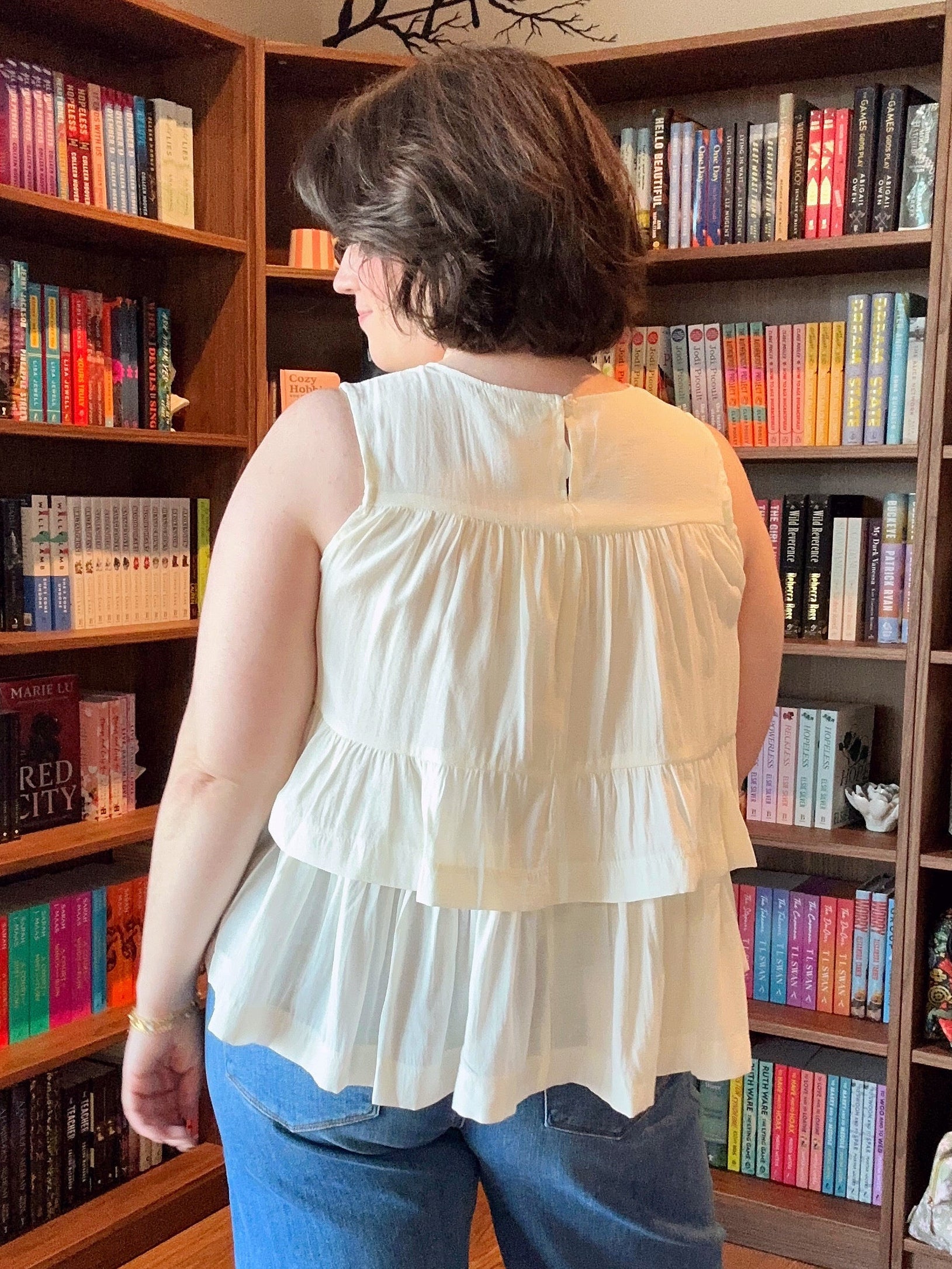 Person wearing a white blouse and blue jeans standing in front of a bookshelf filled with books.
