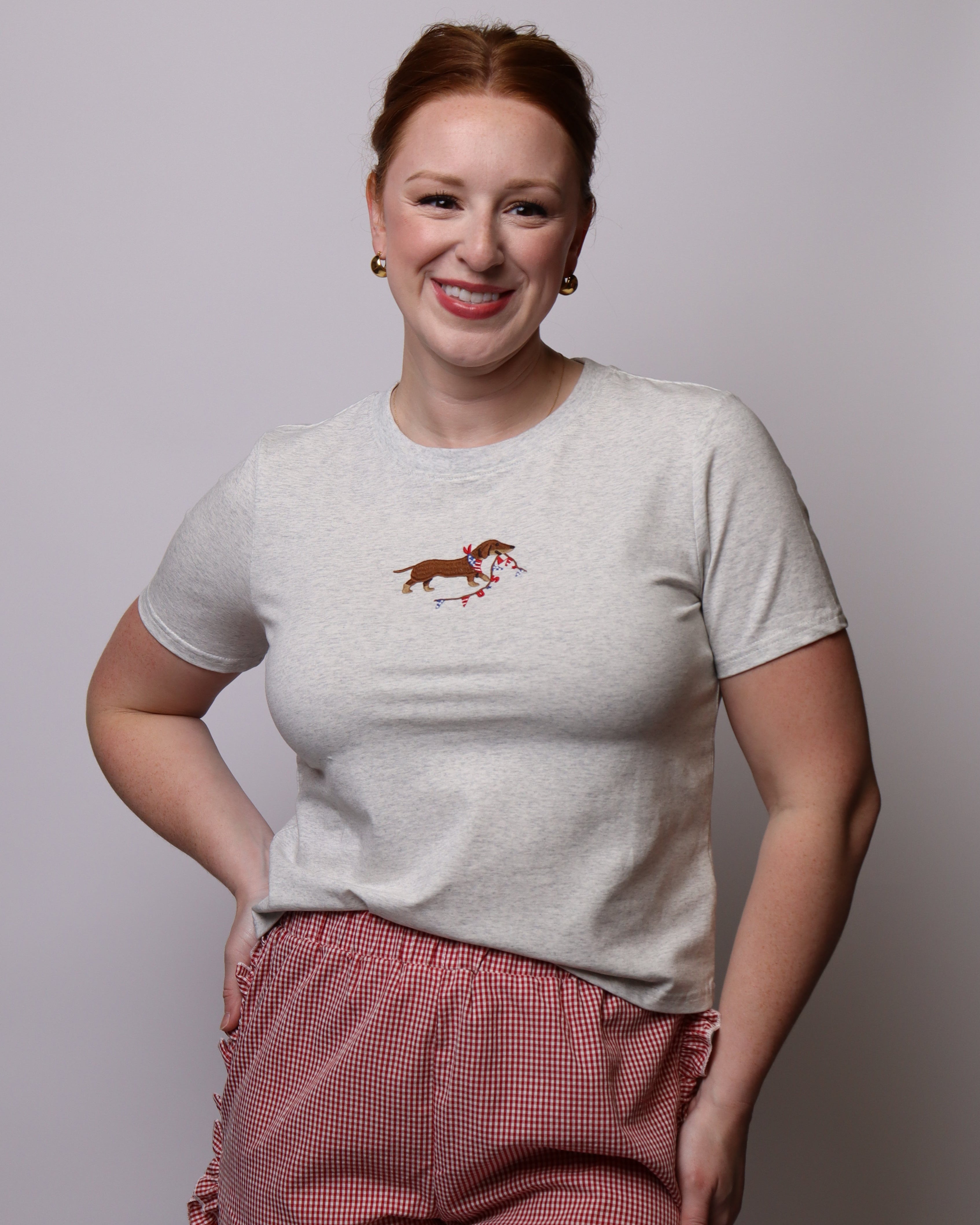 Woman wearing a light gray t-shirt with a embroidered dachshund graphic and red checkered shorts on a plain background