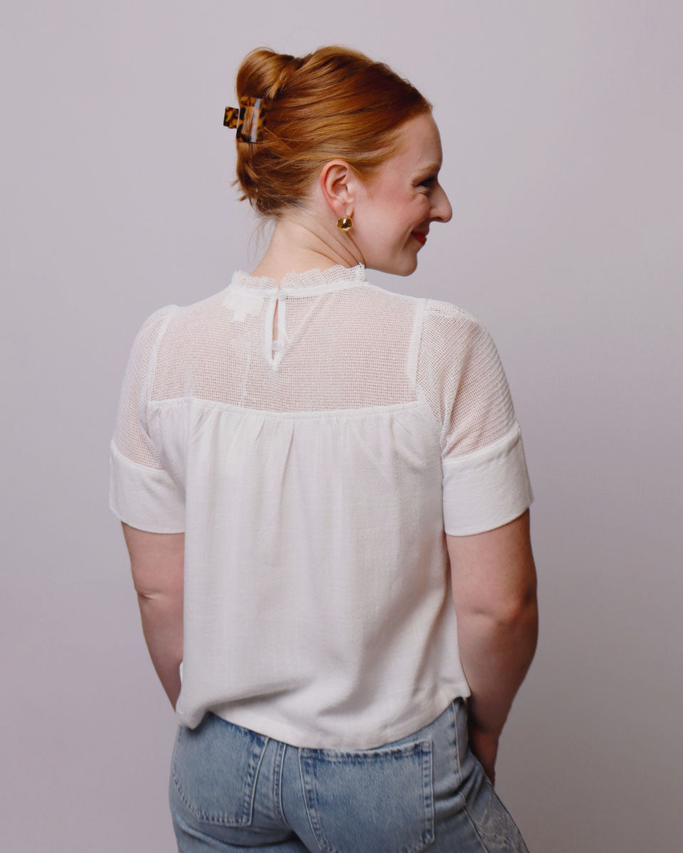 A woman with red hair in a bun is shown from the back wearing a short-sleeve white blouse with sheer mesh back panels and light blue jeans, set against a plain light gray background.