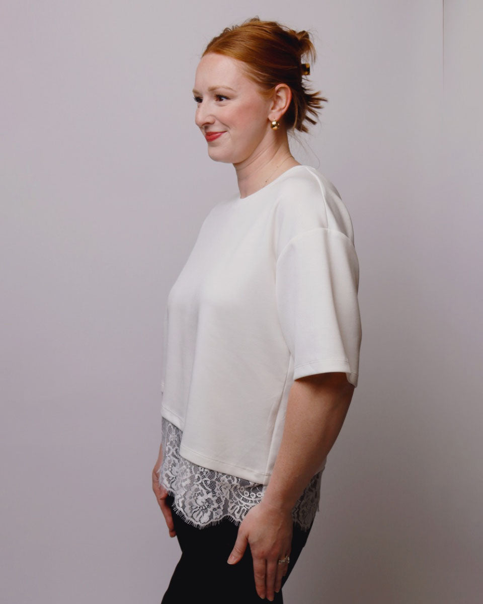 A woman with red hair tied back is shown in profile wearing a white short-sleeve top with a sheer black lace underlayer visible at the hem, paired with black pants, against a plain light gray studio background.