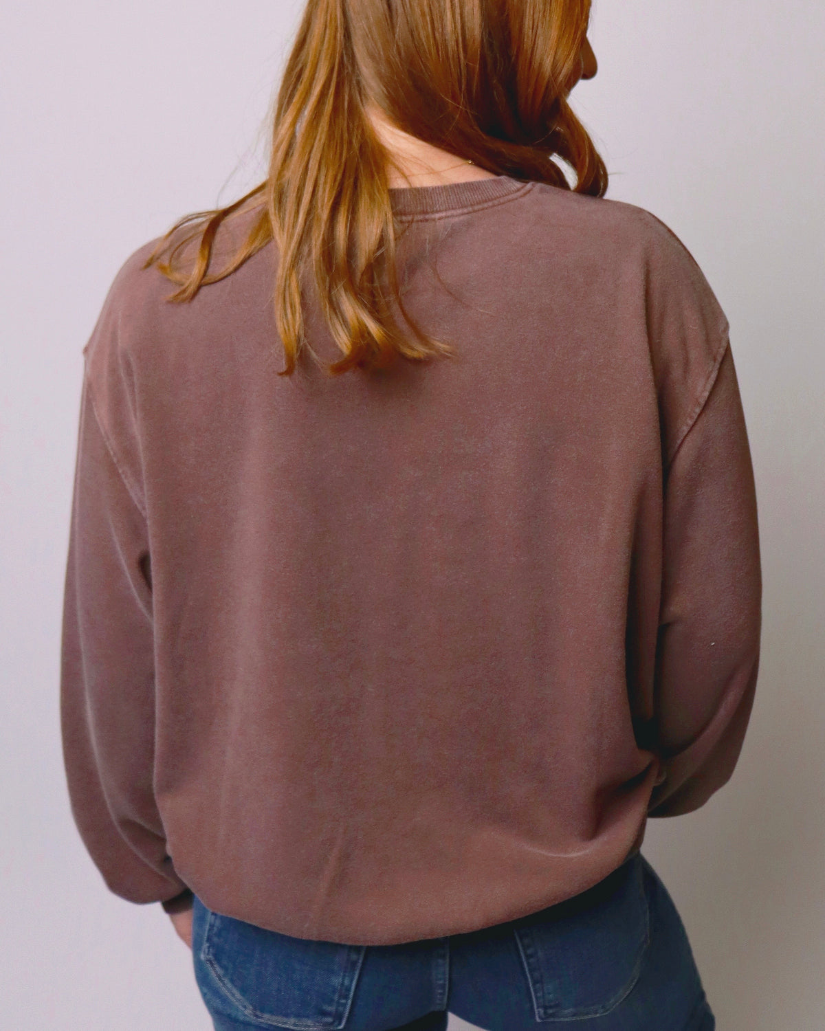 A person viewed from behind wearing a washed, faded brown crewneck sweatshirt and blue jeans against a plain light gray background.
