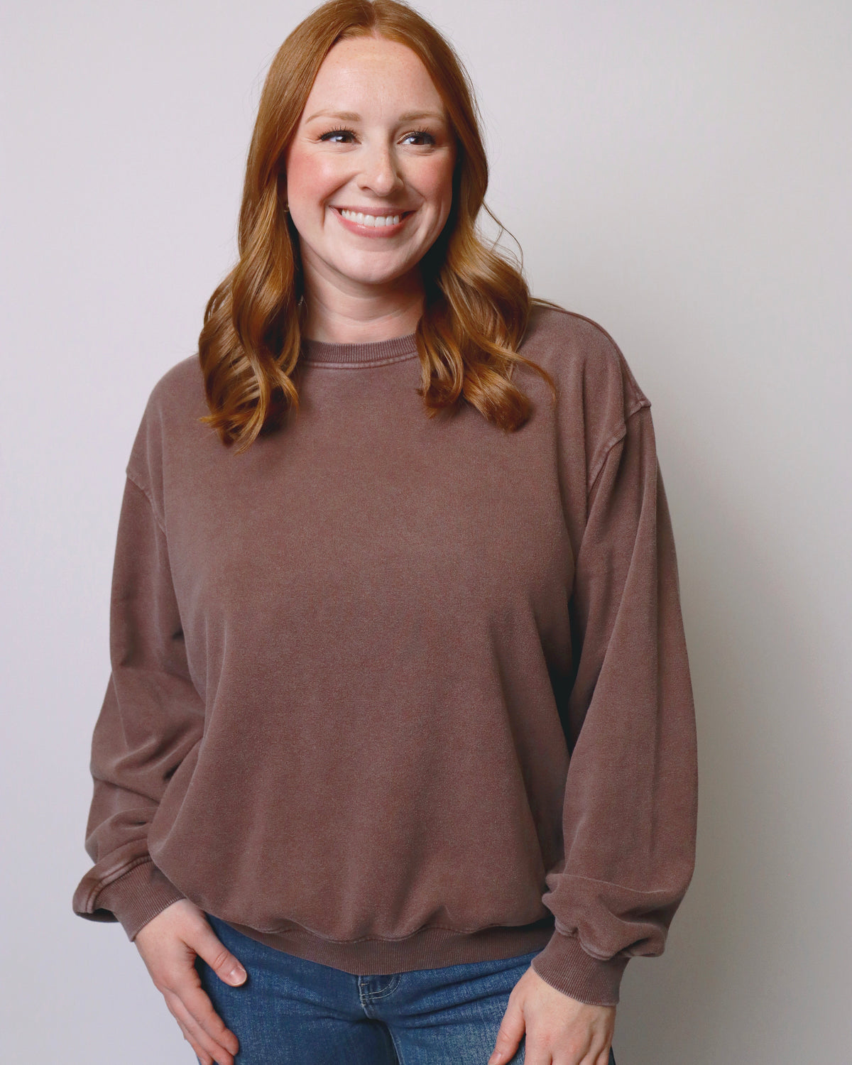 A smiling woman with red hair wearing a brown crewneck sweatshirt and blue jeans, standing against a plain light gray background.