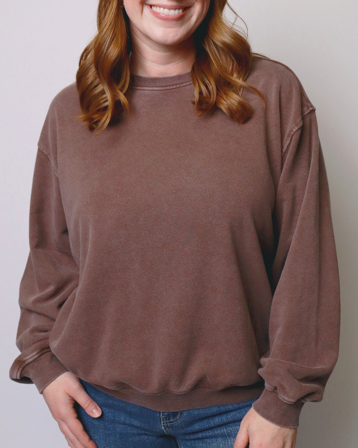 A smiling woman with red hair wearing a brown crewneck sweatshirt and blue jeans, standing against a plain light gray background.