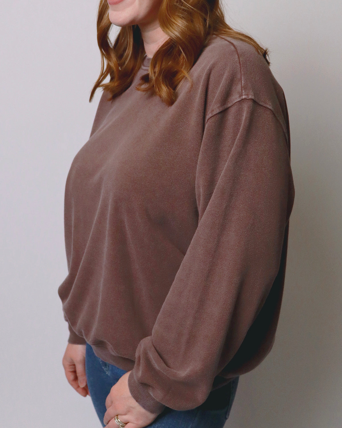 A woman wearing a brown long-sleeve crewneck sweatshirt, shown from the side against a plain light gray background.