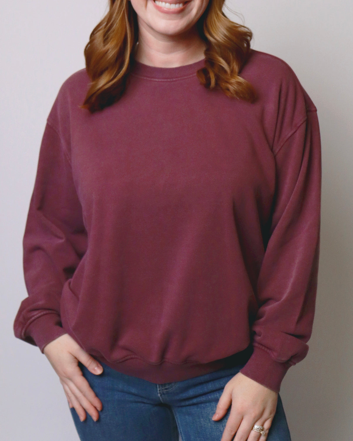 A woman wearing a crewneck sweatshirt in a dusty burgundy color, paired with blue jeans, against a plain light gray background.
