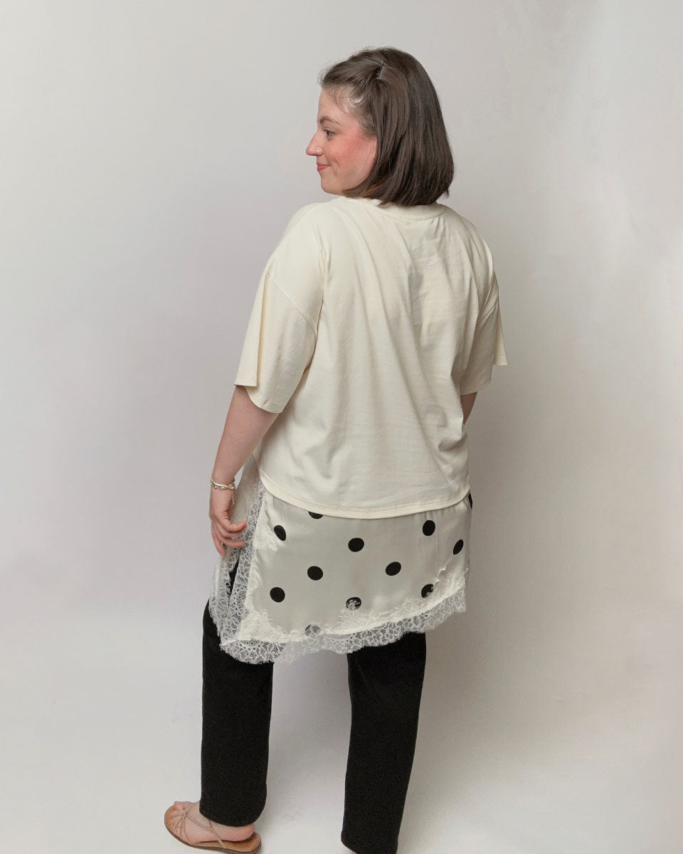 A woman shown from the back wearing a cream-colored short-sleeve top, a white skirt with black polka dots and lace trim, and black pants, standing against a plain light gray studio background.
