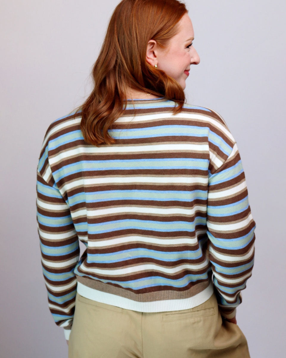 A woman with red hair is shown from the back wearing a long-sleeve knit sweater with horizontal stripes in brown, light blue, and off-white, paired with khaki pants, against a plain light gray background.