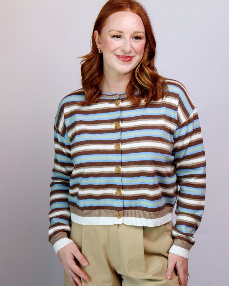 A woman with red hair wearing a cropped, long-sleeve knit cardigan with horizontal brown, beige, and light blue stripes, featuring gold buttons down the front. She is also wearing beige trousers, set against a plain light gray studio background.