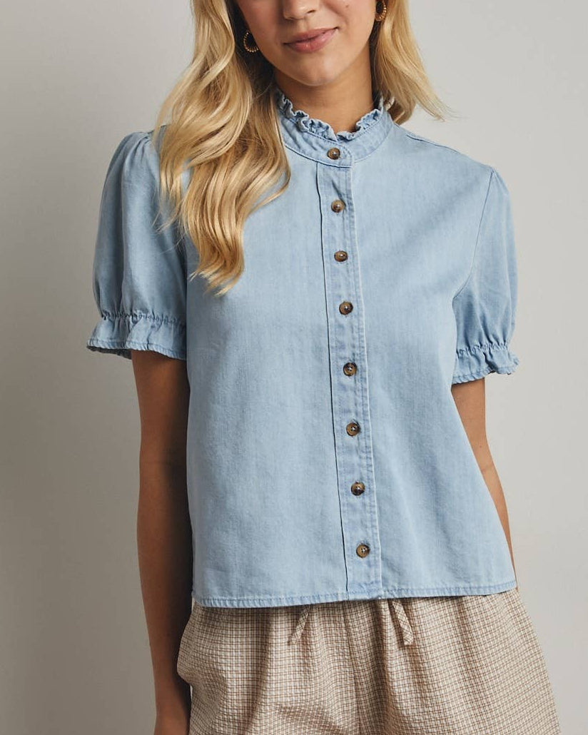 Woman wearing a light blue denim button up shirt with a slight puff sleeve on a neutral background.