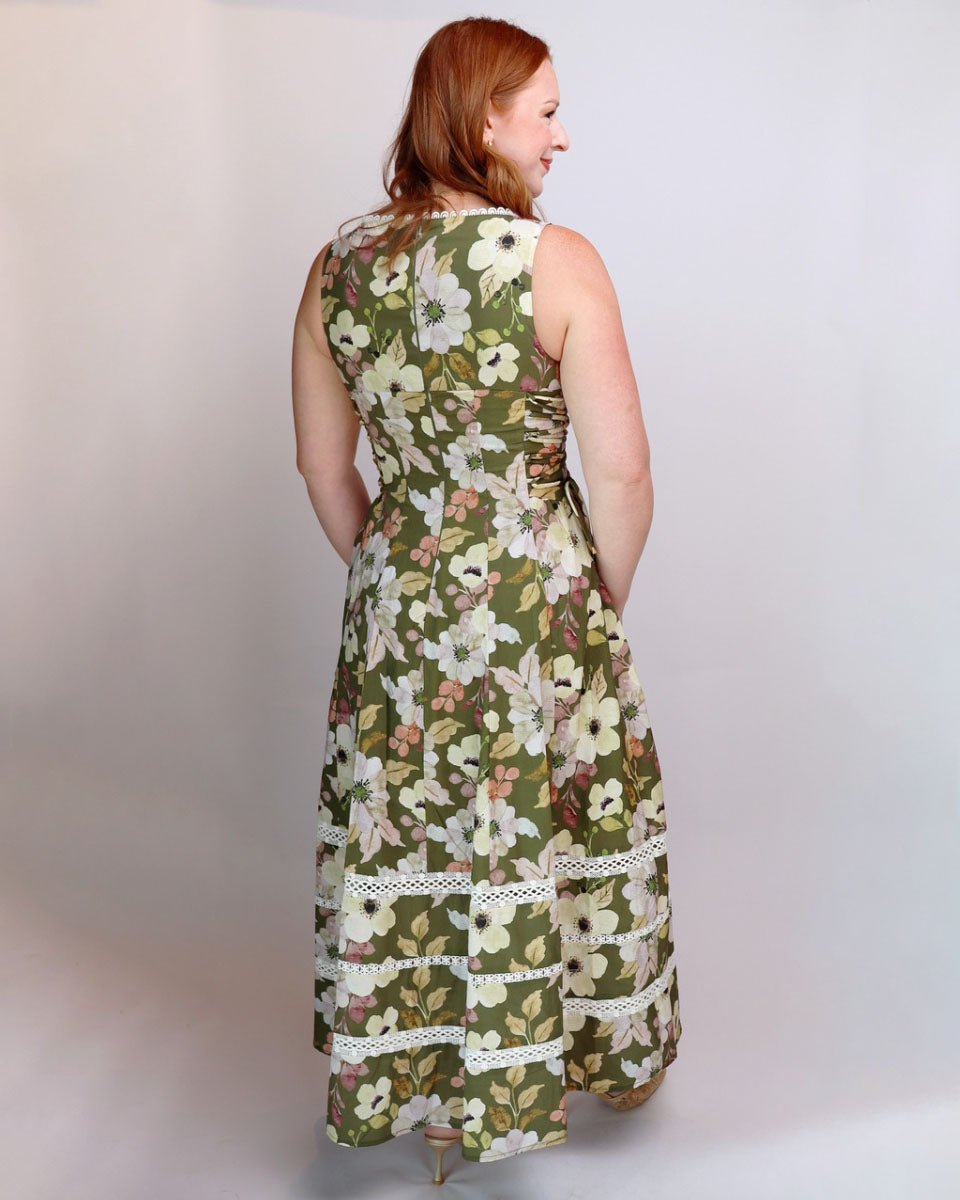 A woman with red hair is shown from the back wearing a sleeveless olive green maxi dress with a white and beige floral pattern and lace trim, set against a plain light gray studio background.