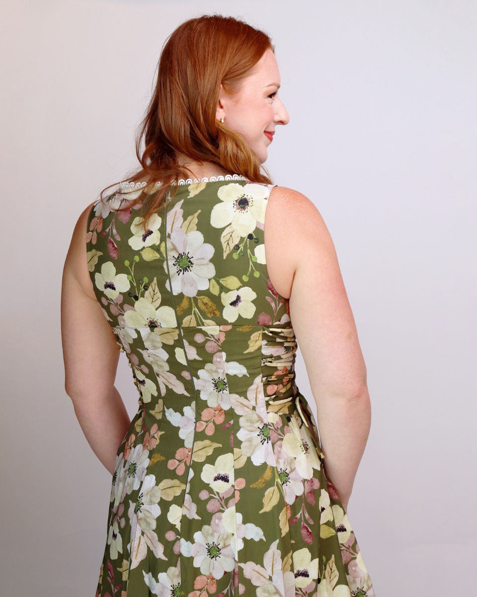 A woman with red hair is shown from the back wearing a sleeveless olive green dress with a large white and beige floral print. She is photographed against a plain light gray studio background.