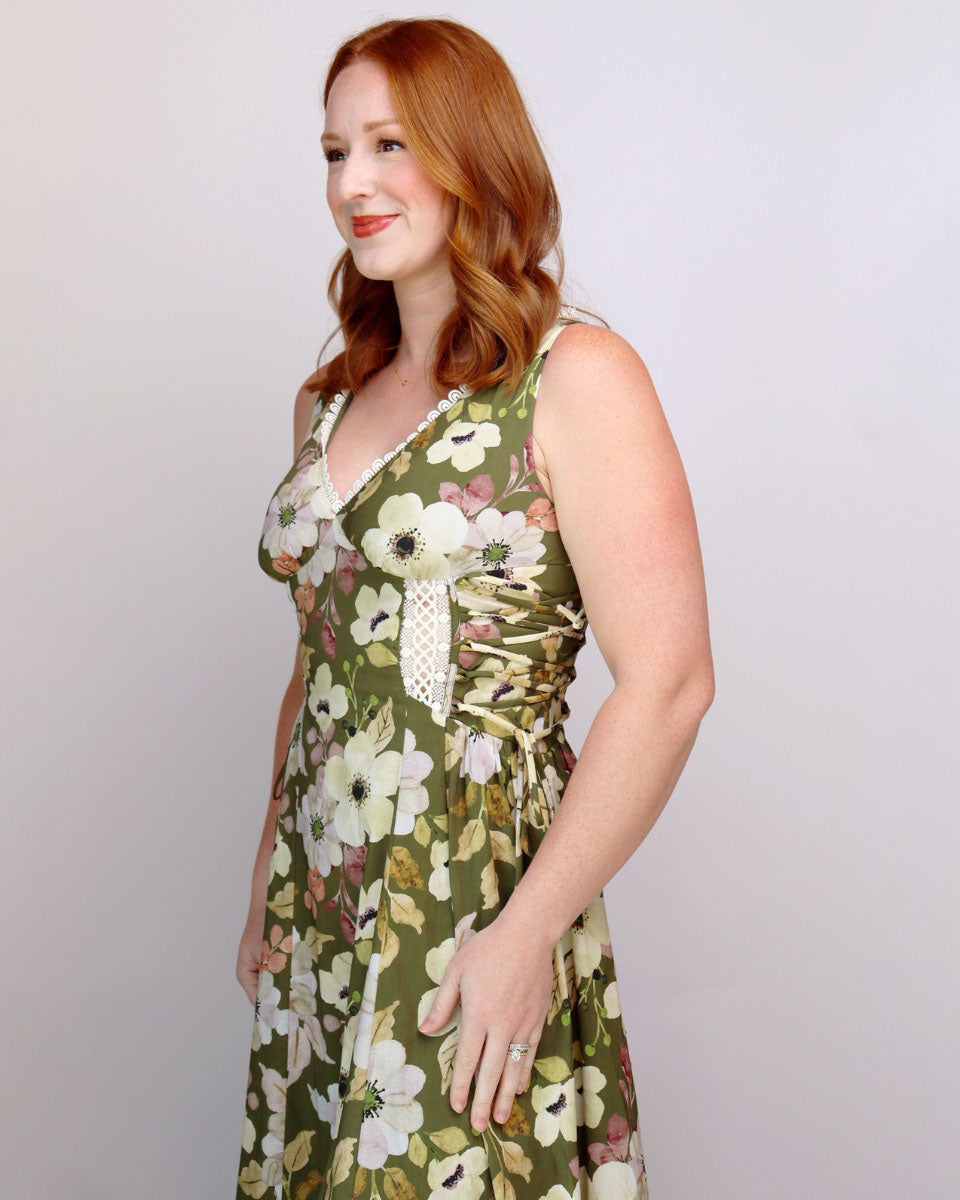 A woman with red hair wearing a sleeveless olive green floral dress with white and pink flowers, standing against a light gray studio background.