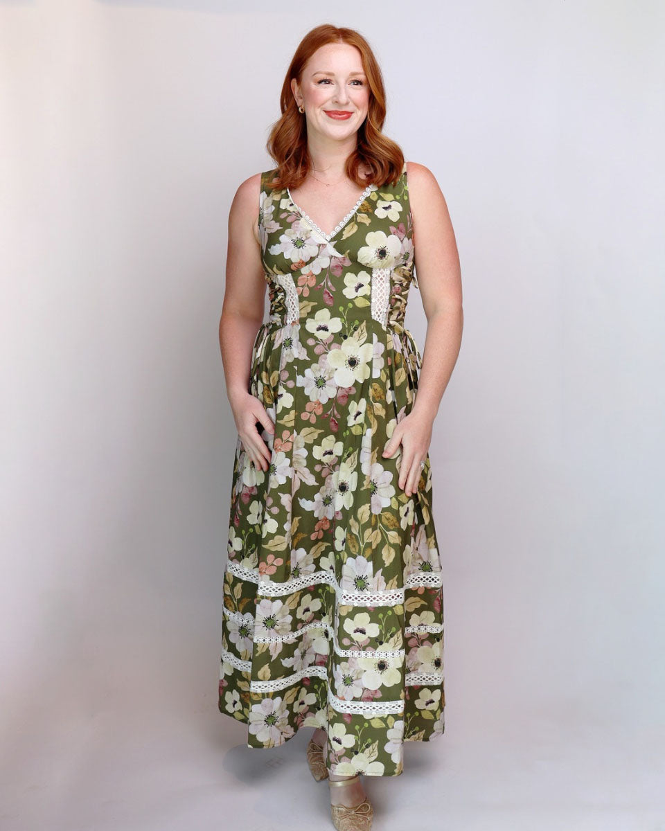A woman with red hair wearing a sleeveless olive green maxi dress with a white and beige floral print and lace trim, standing against a light gray studio background.
