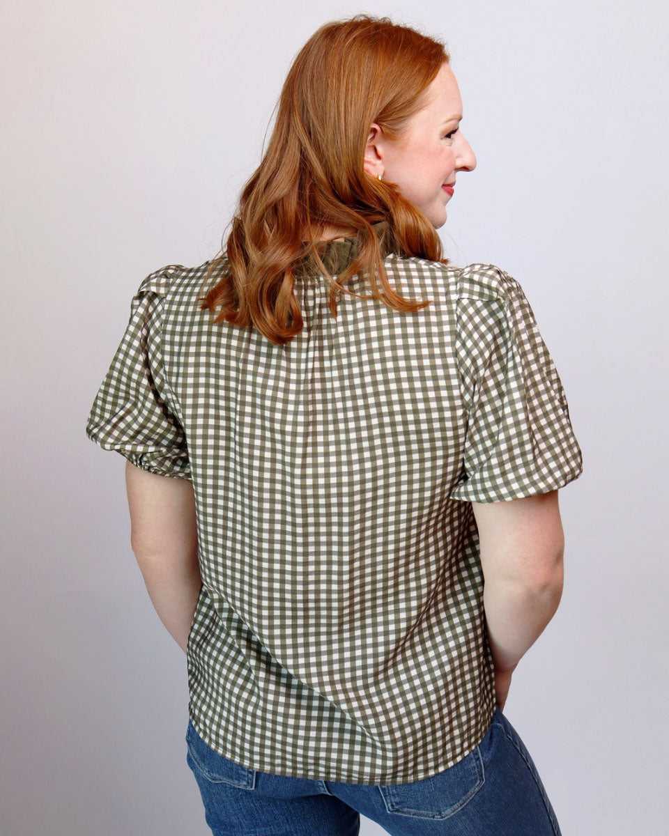 A woman with red hair is shown from the back wearing a short-sleeve gingham check blouse in olive green and white, paired with blue jeans, against a plain light gray background.
