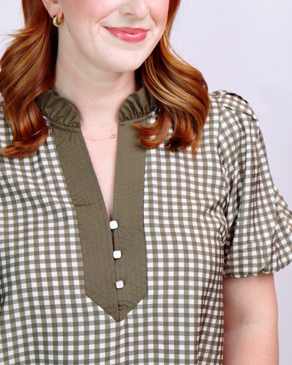 A close-up of a woman wearing a short-sleeve gingham blouse with an olive green ruffled mock neck and button placket.