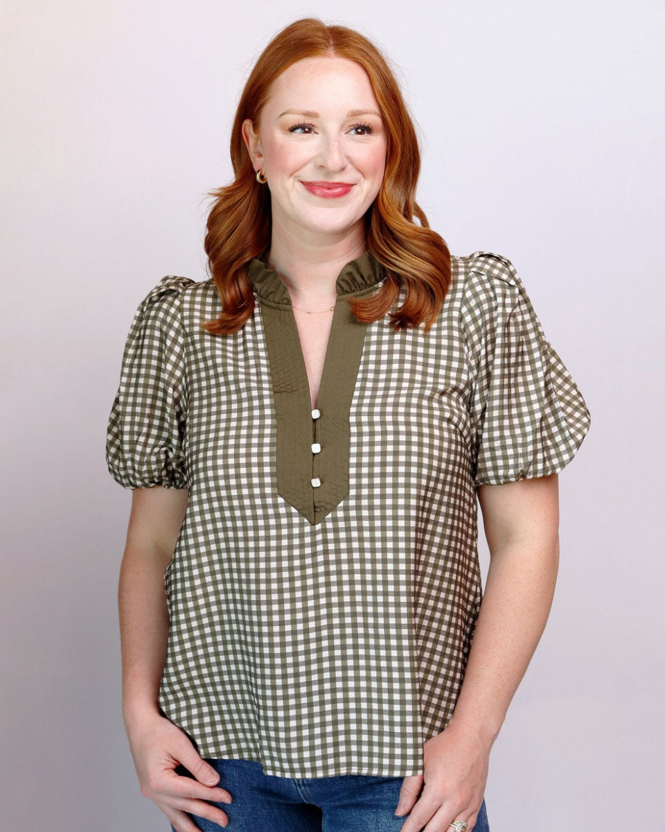 A woman with red hair wearing a short-sleeve gingham check blouse in olive green and white, paired with blue jeans, standing against a light gray studio background.