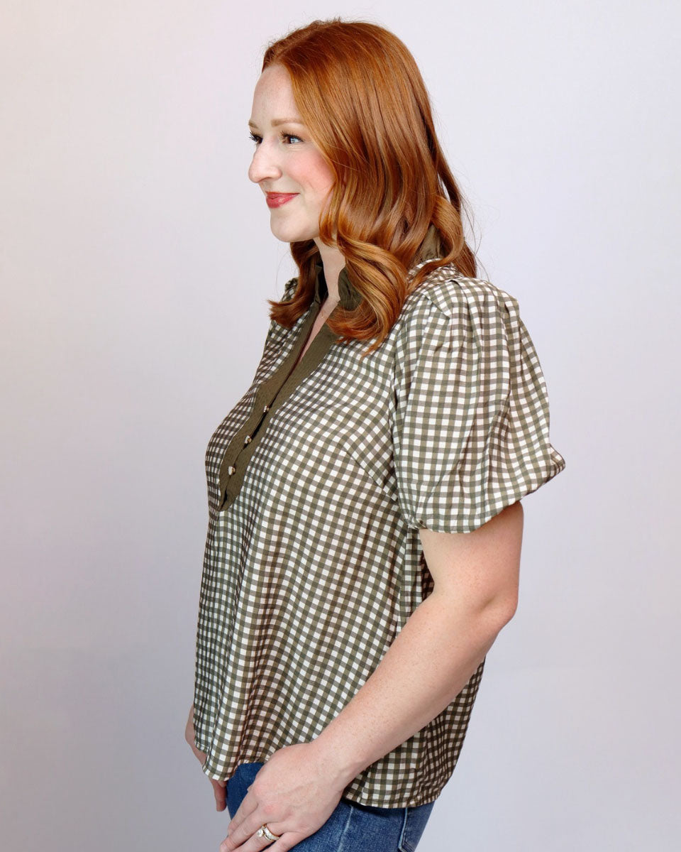 A woman with red hair is shown in profile wearing a short-sleeve gingham blouse in olive green and beige, paired with blue jeans, against a plain light gray studio background.