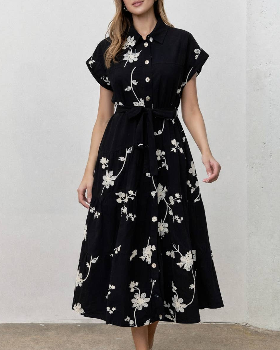 A woman wearing a black midi dress with a white floral print, short cap sleeves, and a matching waist tie, standing against a textured off-white wall.