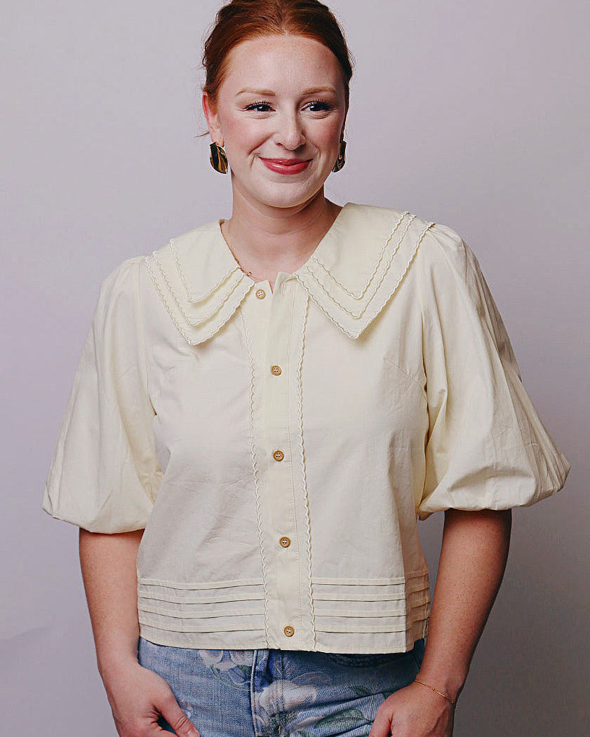 A woman with red hair tied back is wearing a cream-colored short-sleeve blouse with a large decorative collar and button front, paired with blue jeans, against a plain light gray background.