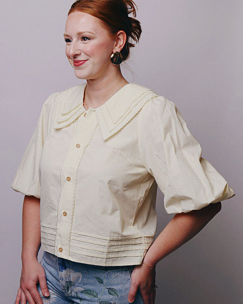 A woman with red hair in an updo wearing a cream-colored short-sleeve blouse with a large collar and puff sleeves, paired with blue floral print jeans, posing against a solid gray studio background.