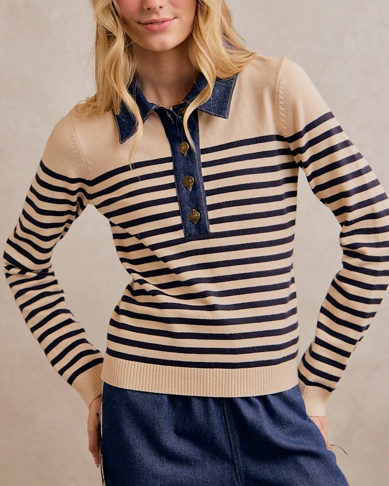 A woman modeling a beige and black striped knit polo sweater with a denim collar and three gold buttons, paired with navy pants, against a neutral beige studio background.
