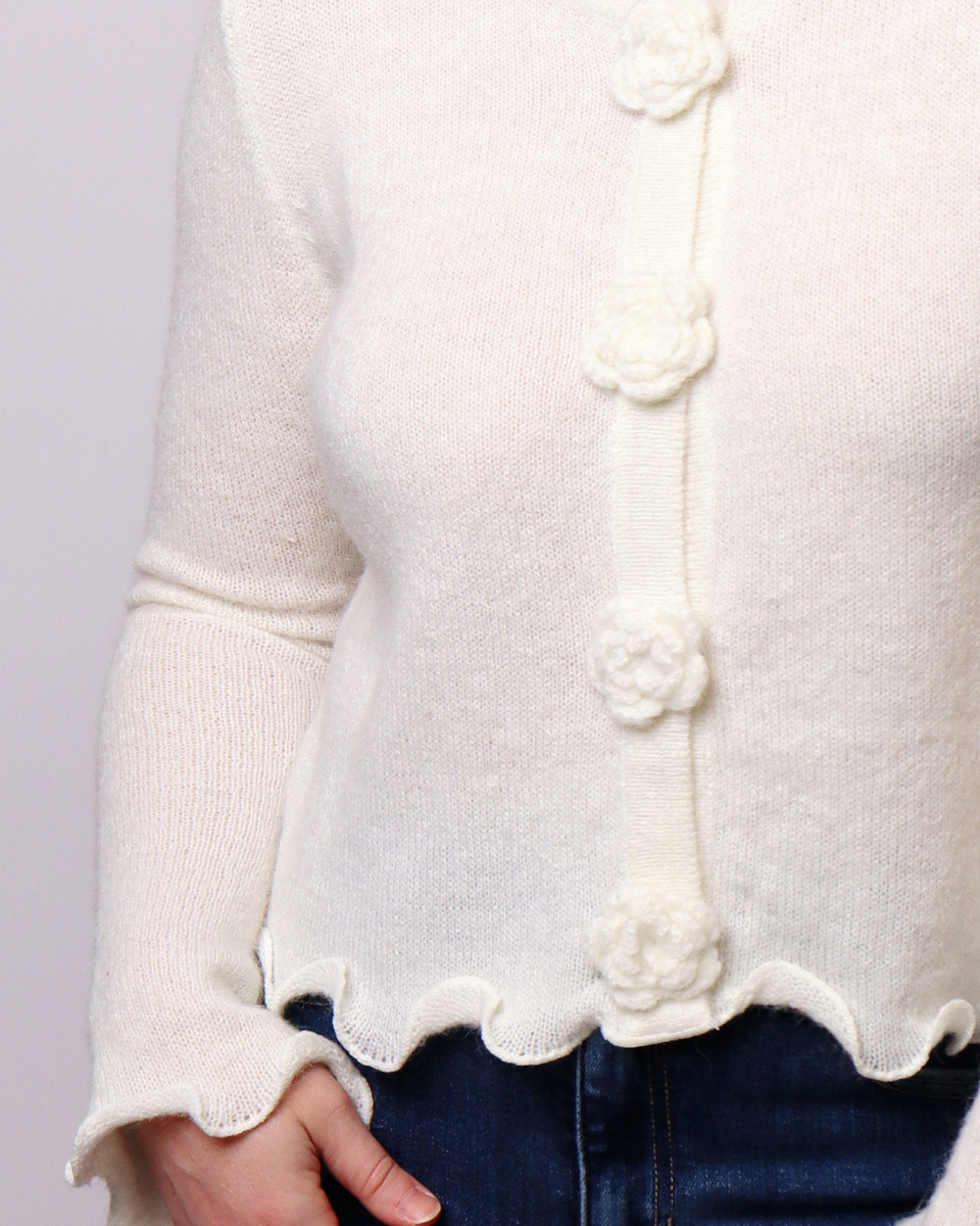Close-up of a cream knit cardigan sweater with textured flower-shaped buttons and a scalloped hem, worn over dark blue jeans.