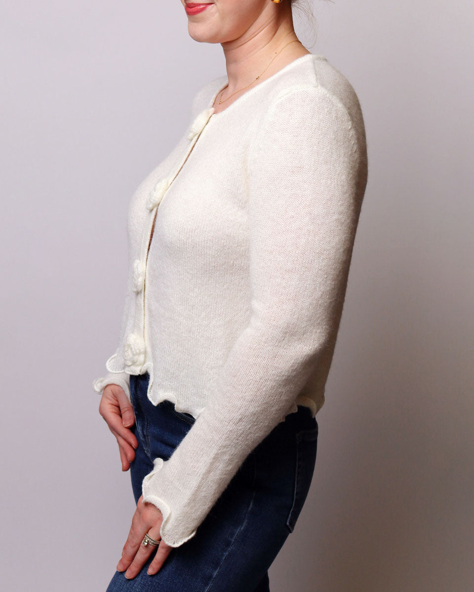 A woman wearing a cream-colored, long-sleeve knit cardigan with decorative buttons, paired with dark blue jeans, shown from a side profile against a plain light gray background.